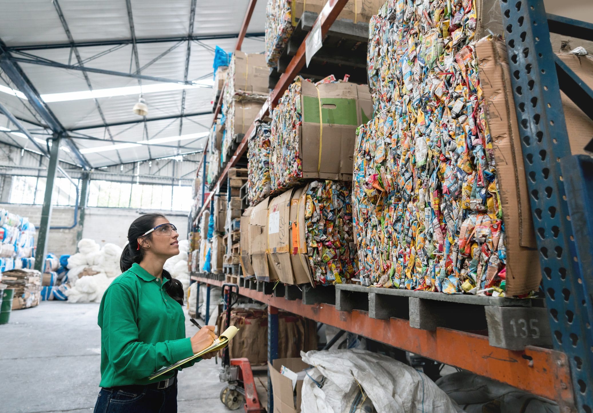 "Une personne en chemise verte avec des lunettes de sécurité inspecte des piles de matériaux recyclables compactés dans un entrepôt