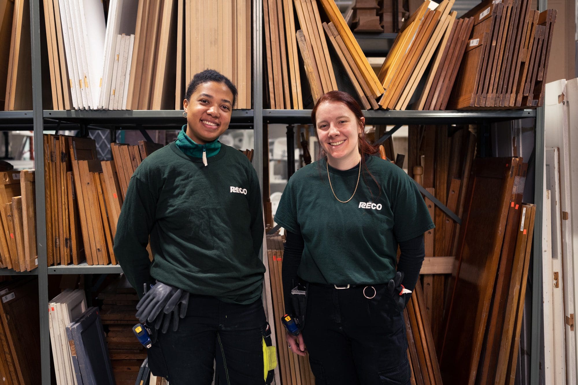 Deux personnes portant des chemises vertes RECCO se tiennent en souriant devant des étagères remplies de diverses planches de bois. Dans ce qui semble être un atelier ou une zone de stockage, l'un tient des gants tandis que l'autre a les mains dans les poches. Ce moment a été capturé par Architectures Sans Frontières.