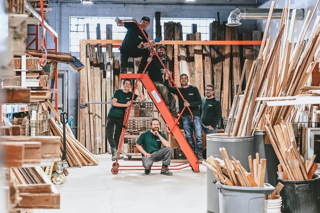 L'équipe d'Architectures Sans Frontières, en tenue décontractée, pose dans un atelier de menuiserie. L'une d'entre elles est assise sur une échelle rouge, incarnant l'esprit de collaboration de l'équipe Architectures Sans Frontières, tandis que les autres se tiennent debout, entourées de planches de bois empilées. L'espace bien éclairé est doté d'une grande fenêtre et d'étagères bien rangées. Crédit photo : Sylviane Robini