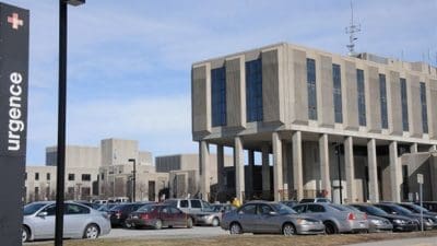 L'image montre un bâtiment hospitalier à l'architecture moderne, doté de grandes fenêtres et soutenu par des piliers. Au premier plan, un parking animé est rempli de voitures. Un panneau indiquant « urgence » marque l'entrée du centre d'urgence de Montréal.