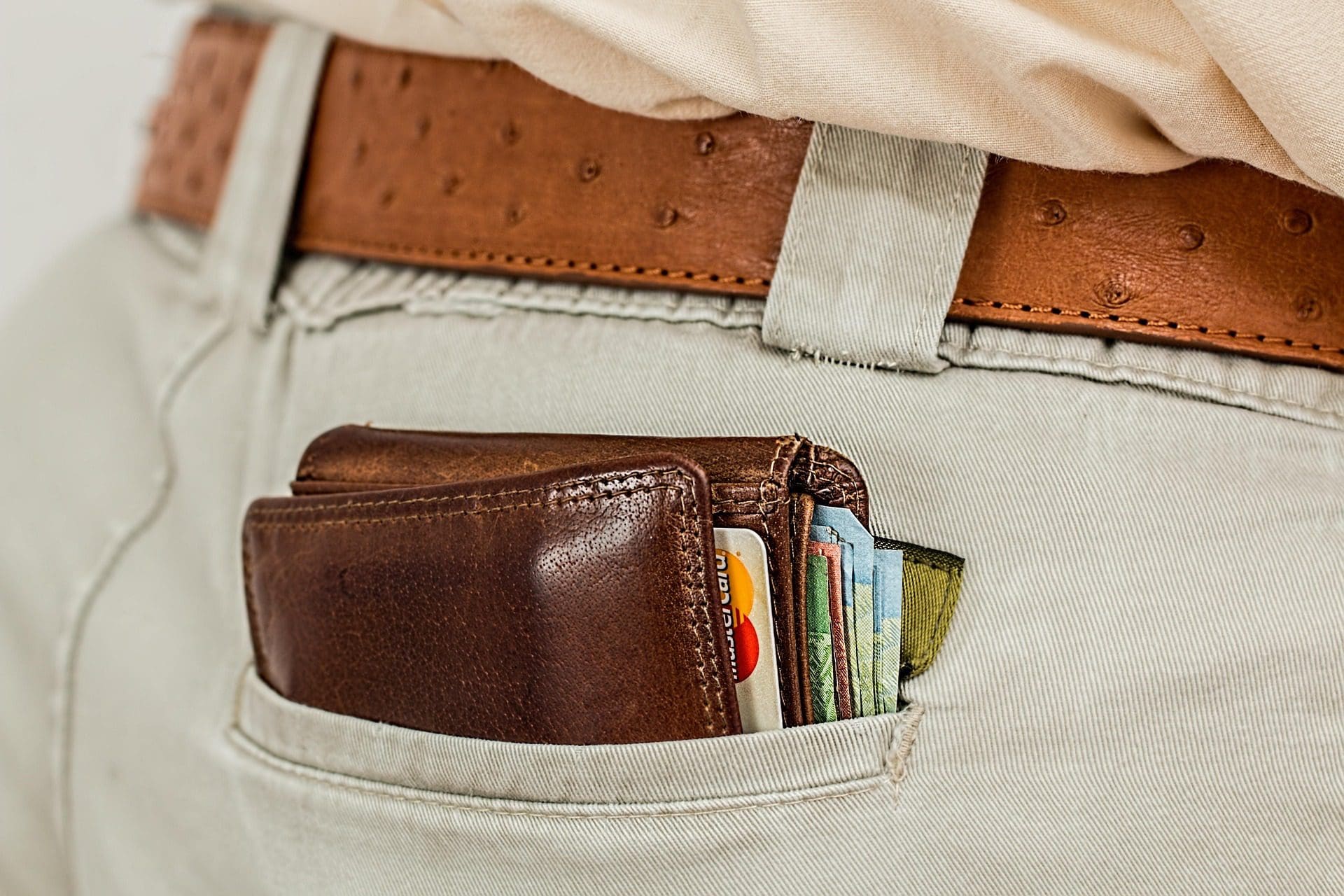 Un portefeuille en cuir marron rempli de cartes et d'argent liquide est partiellement visible dans la poche arrière d'une personne portant un pantalon de couleur claire et une ceinture marron. La poche semble légèrement étirée par le contenu.