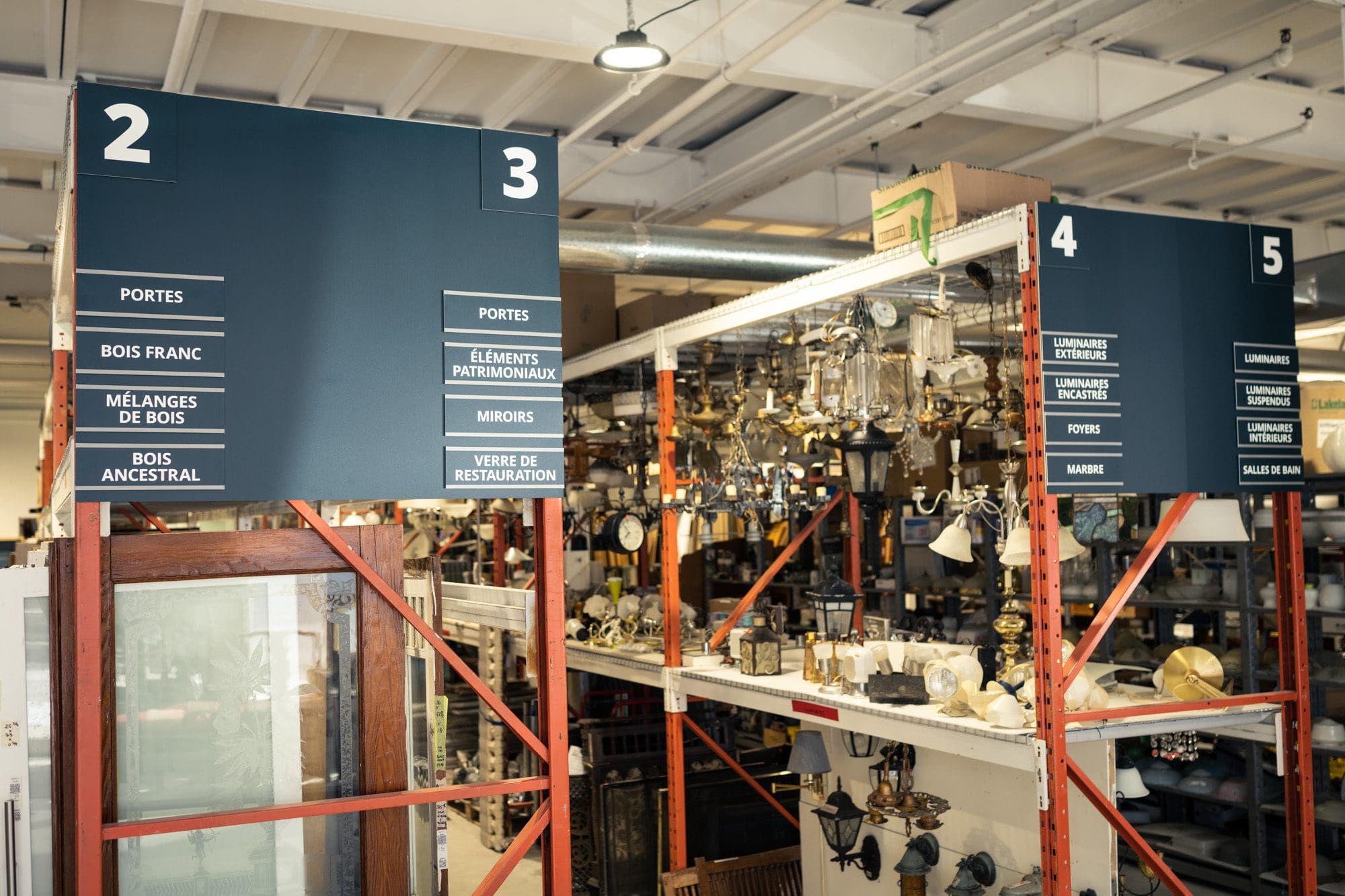 Intérieur d'un entrepôt avec des allées étiquetées. Les panneaux au-dessus des étagères portent les numéros 2 à 5 et répertorient des articles tels que des portes, des bois durs, des miroirs et des appareils d'éclairage. Diverses lampes et quincailleries sont visibles sur les étagères.