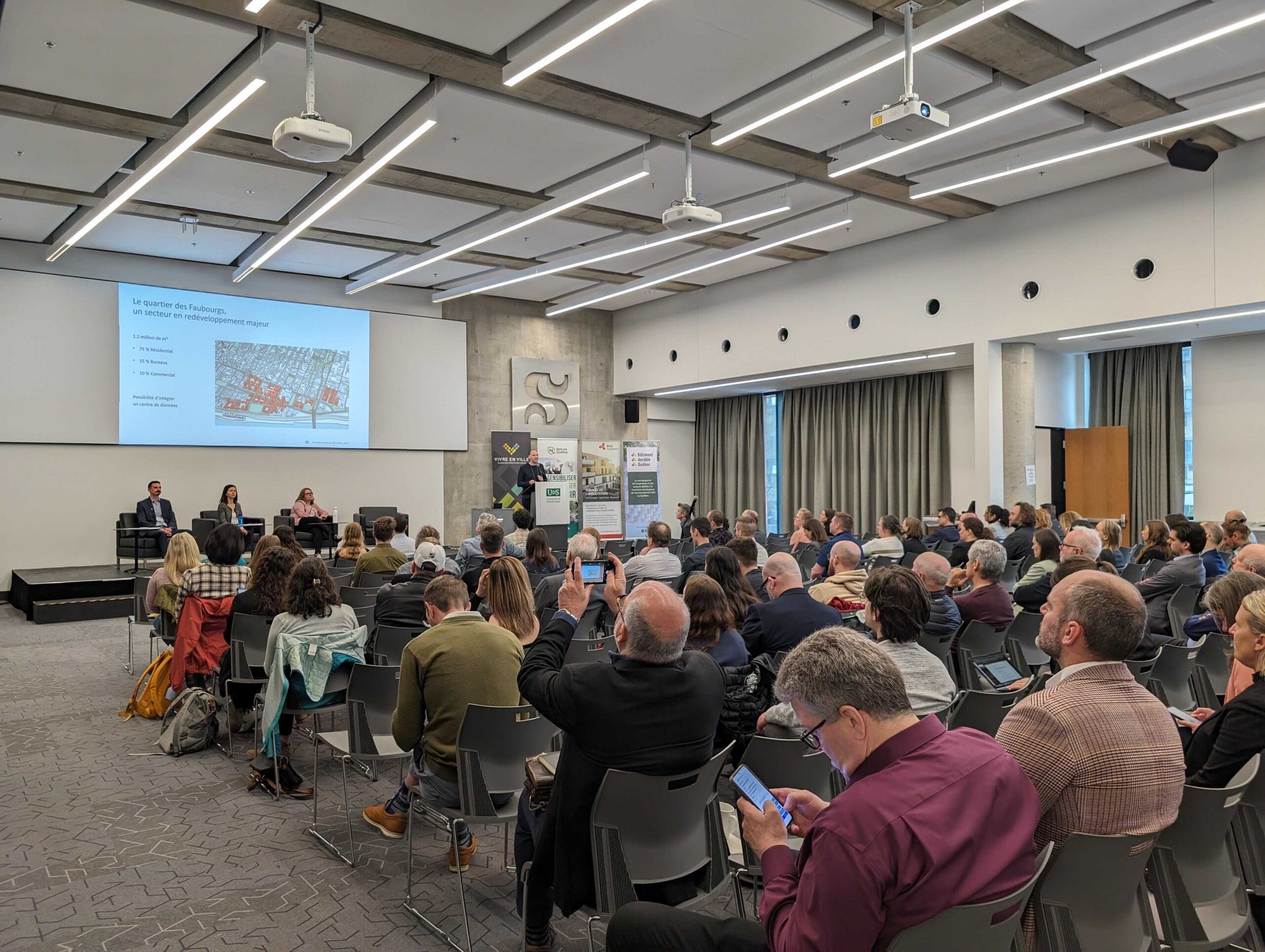 Un large public est assis dans une salle de conférence et suit attentivement une présentation. Trois orateurs se trouvent à l'avant, près d'un écran affichant un schéma architectural. La salle est bien éclairée, avec un design moderne et de hauts plafonds.