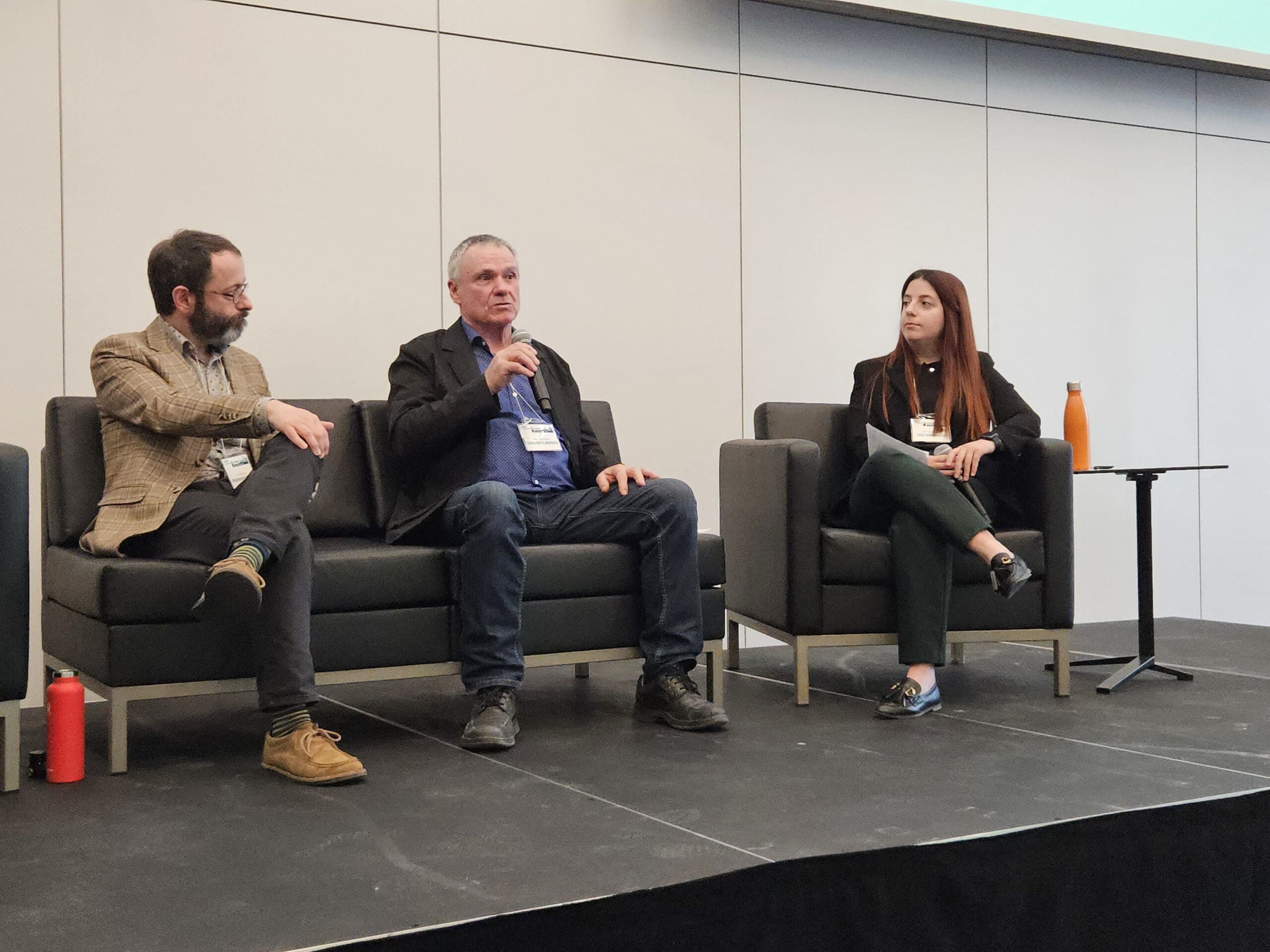 Une personne se tient sur la scène et parle dans un microphone lors du symposium sur les bâtiments sans carbone. Derrière elle, des bannières d'organisations québécoises et un lutrin portant l'inscription UdS Université de Sherbrooke. Le mur de béton est orné d'un logo S.