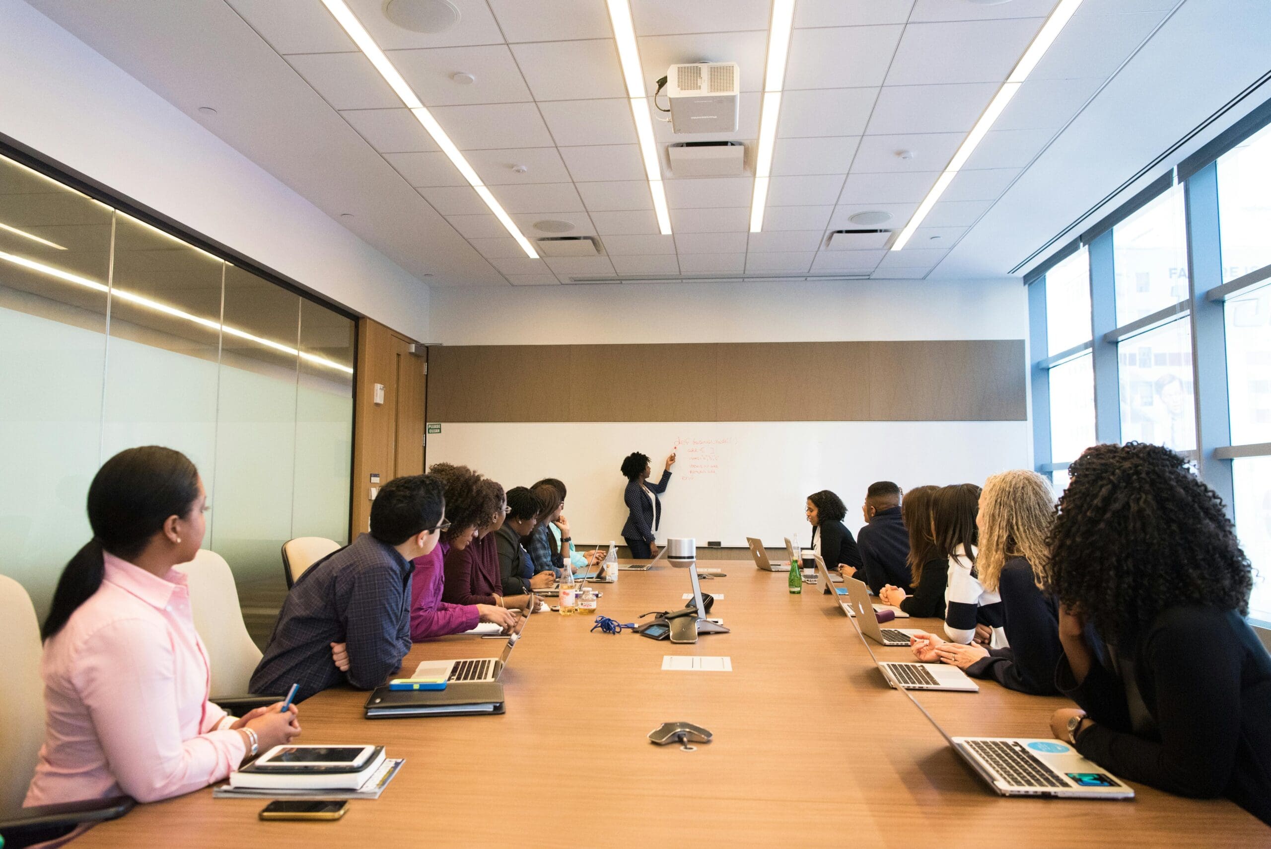 Un groupe diversifié de personnes en tenue professionnelle est réuni autour d'une table de conférence dans un bureau moderne. Elles regardent attentivement quelqu'un présenter les détails de la boucle énergétique de Saint-Laurent sur un tableau blanc, avec des ordinateurs portables et des cahiers répartis sur la table.