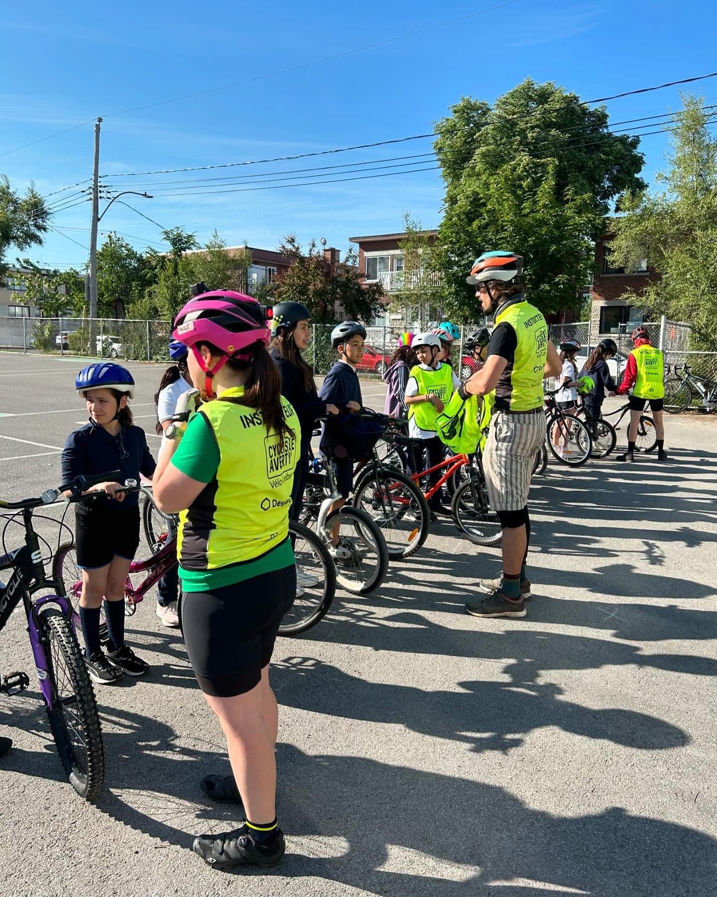 Un groupe de personnes portant des casques, dans le cadre du programme Cycliste Averti de Vélo Québec, se tient avec leurs vélos sur une aire pavée. La plupart d'entre eux portent des maillots cyclistes verts. La scène est ensoleillée, les arbres et les maisons forment une charmante toile de fond.
