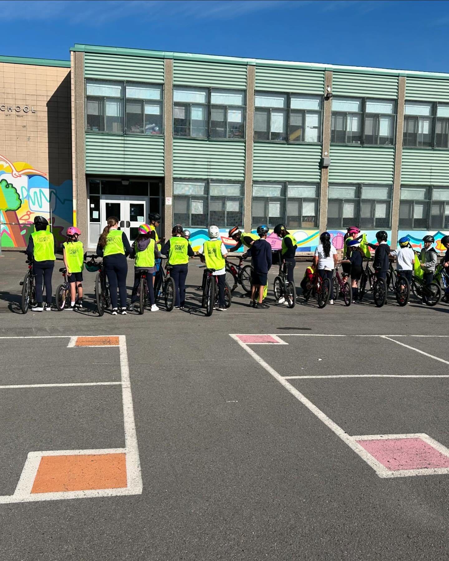 Un groupe de personnes portant des casques et des gilets fluo se tient avec des vélos devant un bâtiment scolaire, participant au programme Cycliste Averti de Vélo Québec. Le bâtiment est orné d'une peinture murale colorée et de grandes fenêtres, tandis que le sol présente des carrés peints sous un ciel clair et ensoleillé.