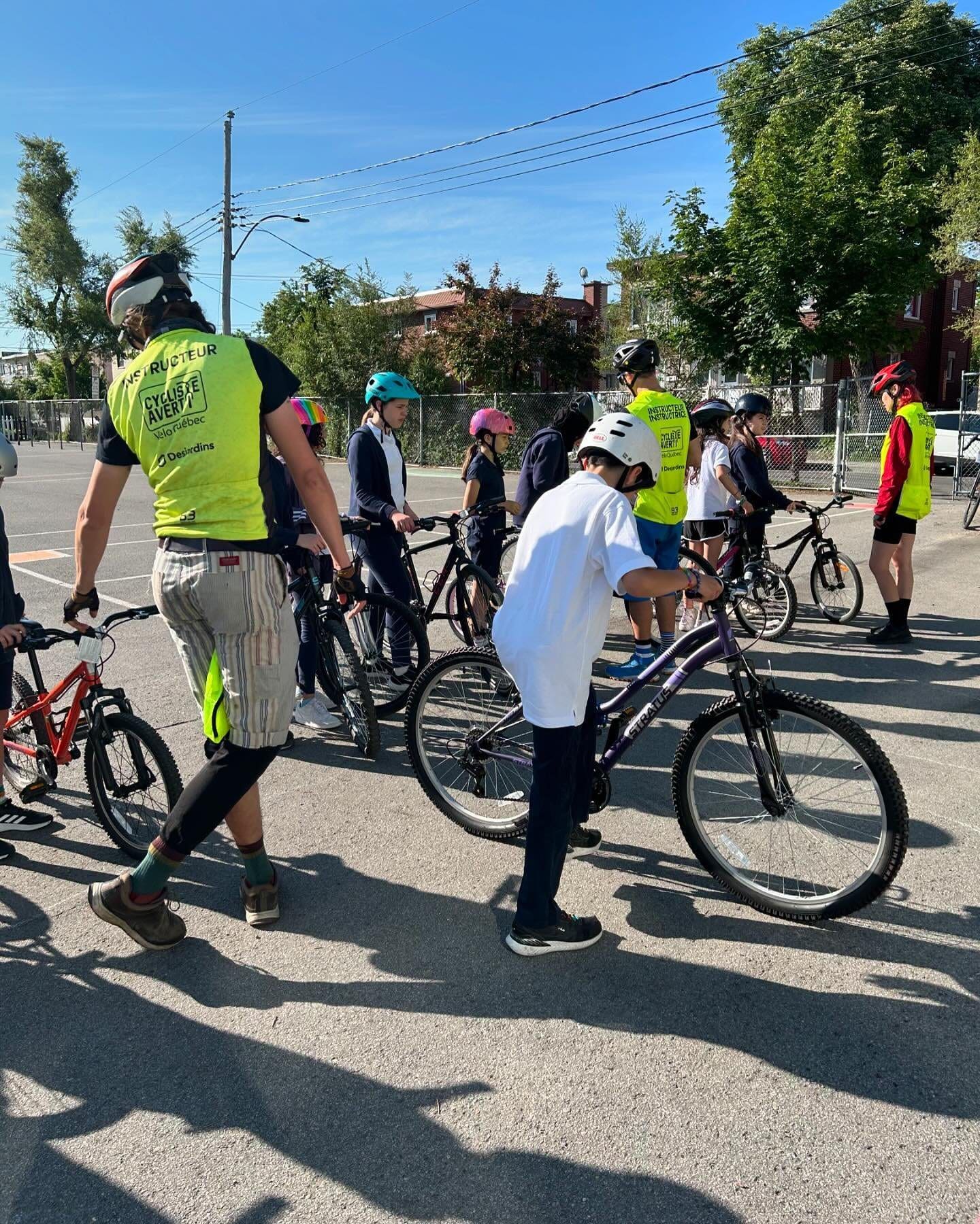 Un groupe de cyclistes, des adultes aux enfants, se réunit par une journée ensoleillée avec des vélos. Les participants au programme Cycliste Averti de Vélo Québec portent fièrement leur casque et leur équipement cycliste. Des arbres et des bâtiments encadrent la scène animée de cet événement cycliste communautaire dynamique.