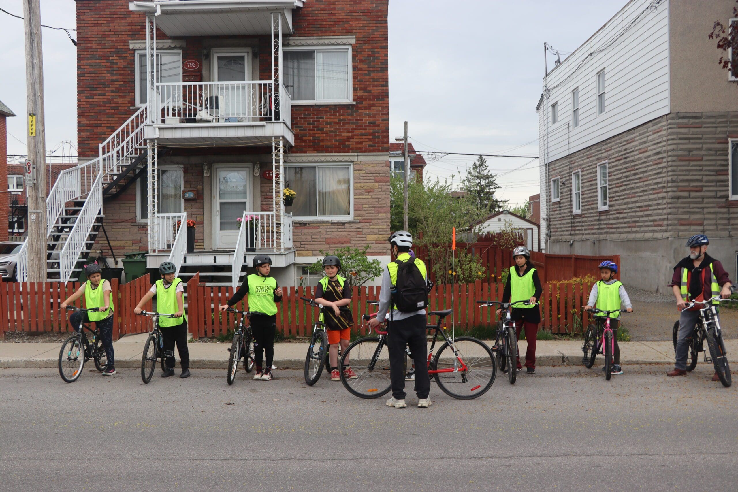 Un groupe de cyclistes, vêtus de gilets de sécurité de couleur vive, se rassemble dans une rue résidentielle dans le cadre du programme Cycliste Averti de Vélo Québec. Un instructeur, également vêtu d'un gilet, se tient debout, un vélo face à eux. Des maisons en briques avec des escaliers et une clôture rouge forment la toile de fond.