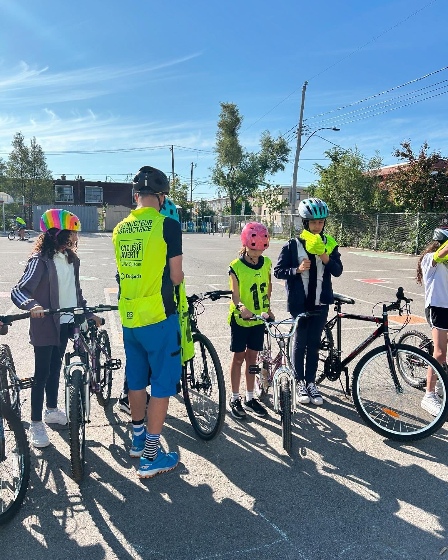 Un groupe d'enfants et un adulte portant des casques et des gilets de sécurité brillants se tiennent près de leurs bicyclettes sur une aire pavée, participant au programme Cycliste Averti de Vélo Québec. L'adulte semble donner des instructions aux enfants. Le ciel est dégagé, les arbres et les bâtiments sont visibles en arrière-plan.