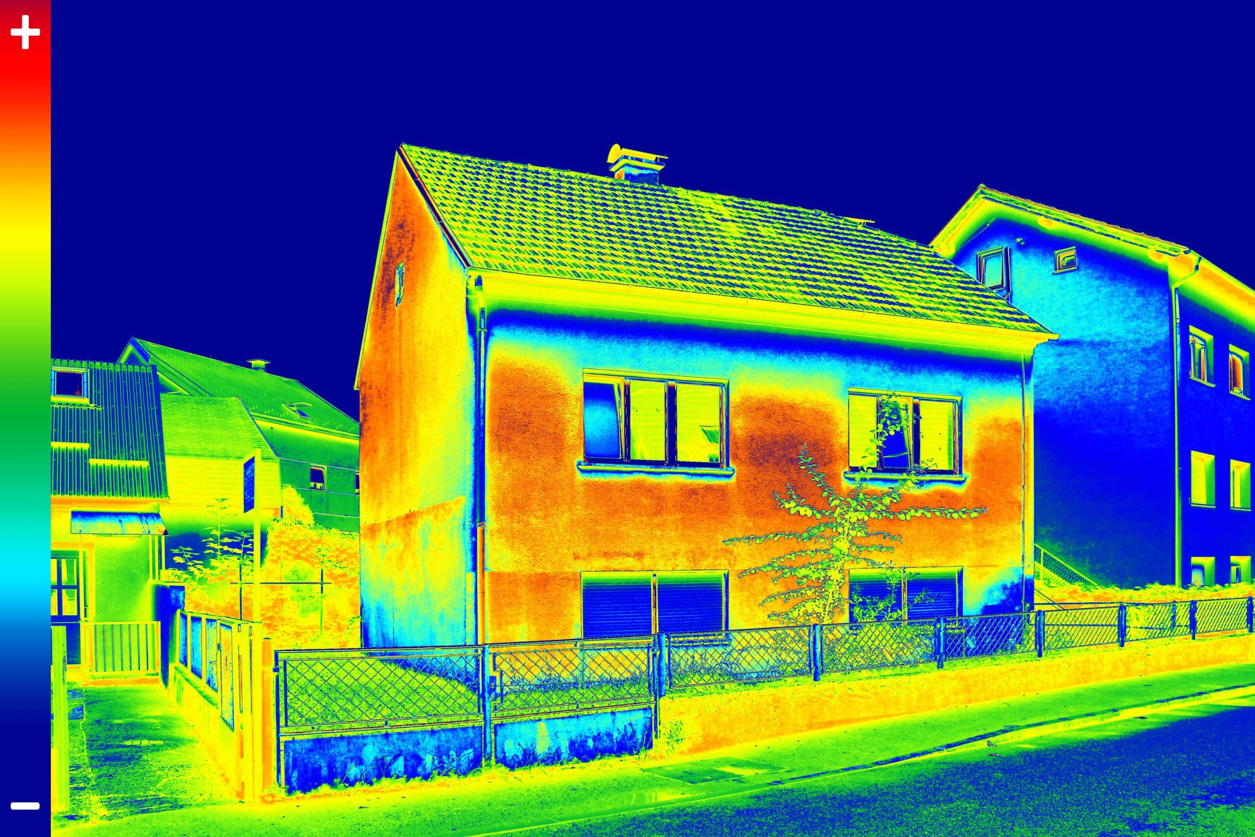 Une image thermique d'une rue résidentielle montre une maison à deux étages avec des variations de température. La maison est principalement rouge et orange, ce qui indique des zones plus chaudes, avec des teintes bleues et vertes plus froides sur le toit et les bâtiments voisins.