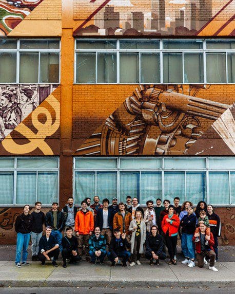 Un groupe de personnes, connu sous le nom d'équipe de la Coop Centrale Agricole, se tient serré devant un bâtiment orné d'une grande peinture murale colorée représentant des motifs abstraits et des éléments mécaniques. Certains portent des vestes et un chien est assis à l'avant.