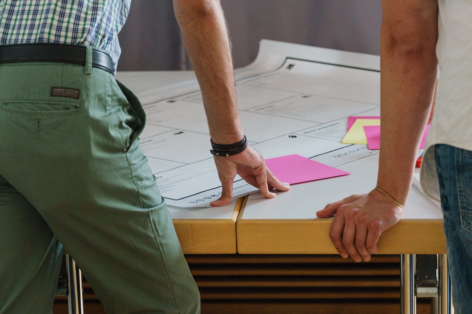 Deux personnes sont accoudées à une table et examinent de grandes feuilles de papier ou des plans. L'une porte un pantalon vert et une chemise à carreaux, tandis que l'autre est vêtue d'un tee-shirt blanc. Des notes autocollantes roses sont apposées sur les feuilles.
