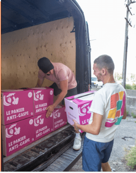 Deux personnes chargent des cartons étiquetés Le Panier Anti-Gaspi à l'arrière d'une camionnette. Une personne, portant un bonnet et une chemise rose, se trouve à l'intérieur de la camionnette, tandis que l'autre, vêtue d'une chemise multicolore, se tient à l'extérieur, un carton à la main.