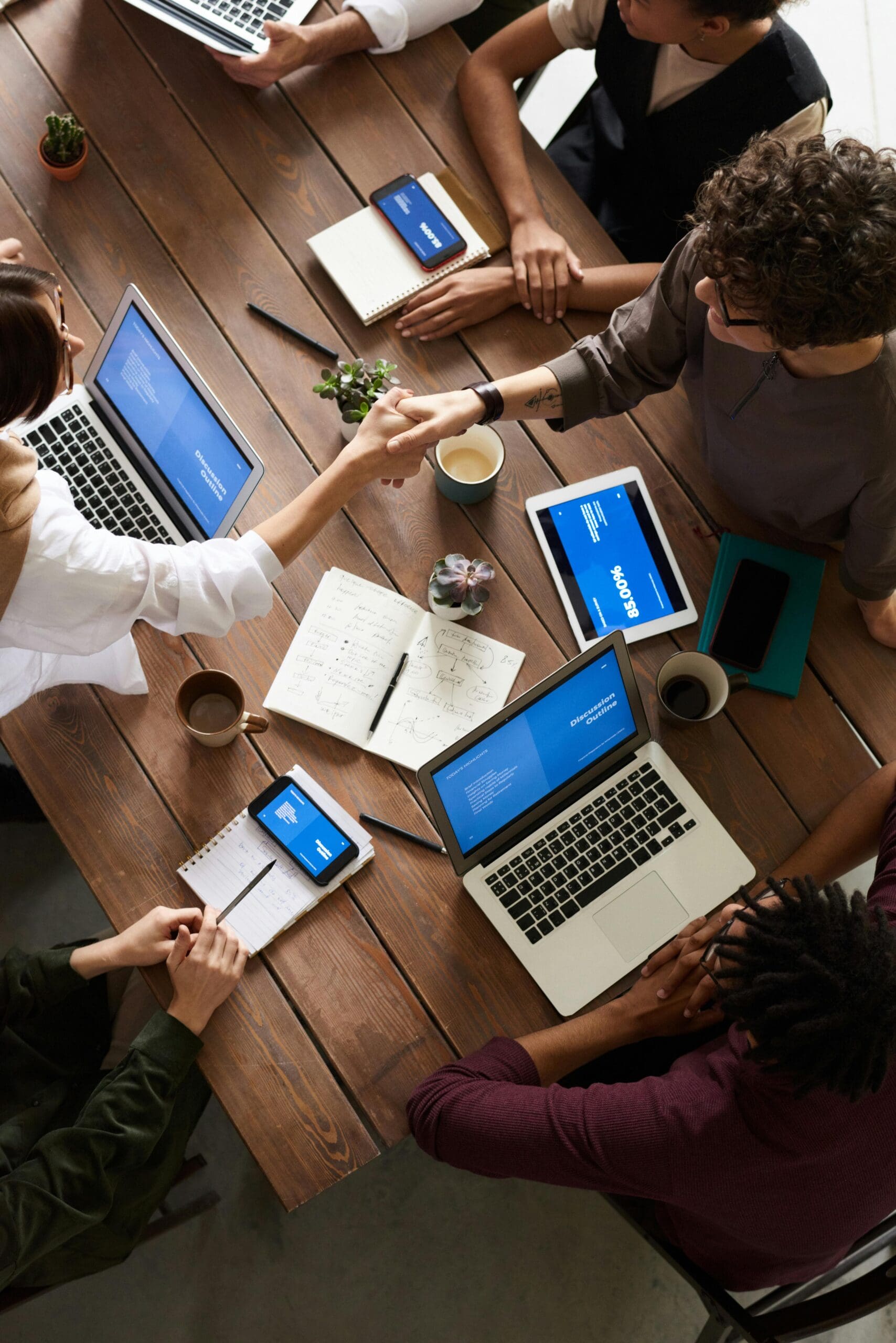 Un groupe de personnes assises autour d'une table en bois avec des ordinateurs portables, des tablettes, des carnets de notes et des tasses de café. Elles semblent être en réunion, en train de discuter ou de collaborer, certaines se serrant la main. Tous les écrans affichent des graphiques bleus similaires.