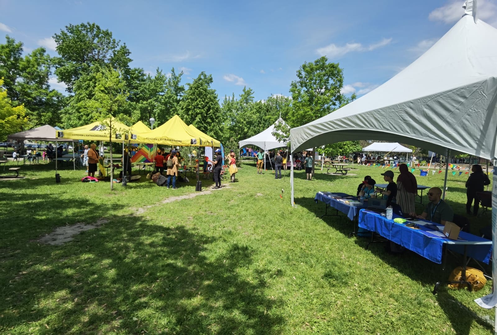 Un événement MobiliSENS en plein air et ensoleillé se déroule sous des tentes blanches et jaunes qui parsèment l'espace herbeux. Les gens se rassemblent près des tentes, profitant des tables installées sous les couvertures. Des arbres luxuriants et un ciel d'un bleu éclatant offrent une toile de fond pittoresque à cette scène animée.