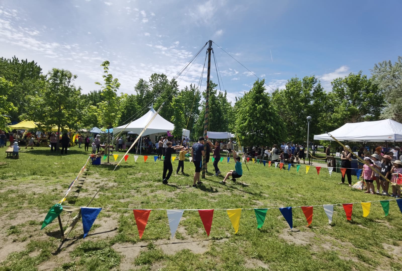 L'événement MobiliSENS transforme un parc parsemé d'arbres verts en un festival en plein air très animé. Les gens se rassemblent autour de tentes et de stands animés, tandis que des drapeaux colorés délimitent une zone centrale où se déroulent des spectacles. Le ciel est partiellement nuageux, révélant des taches de bleu au milieu des festivités.