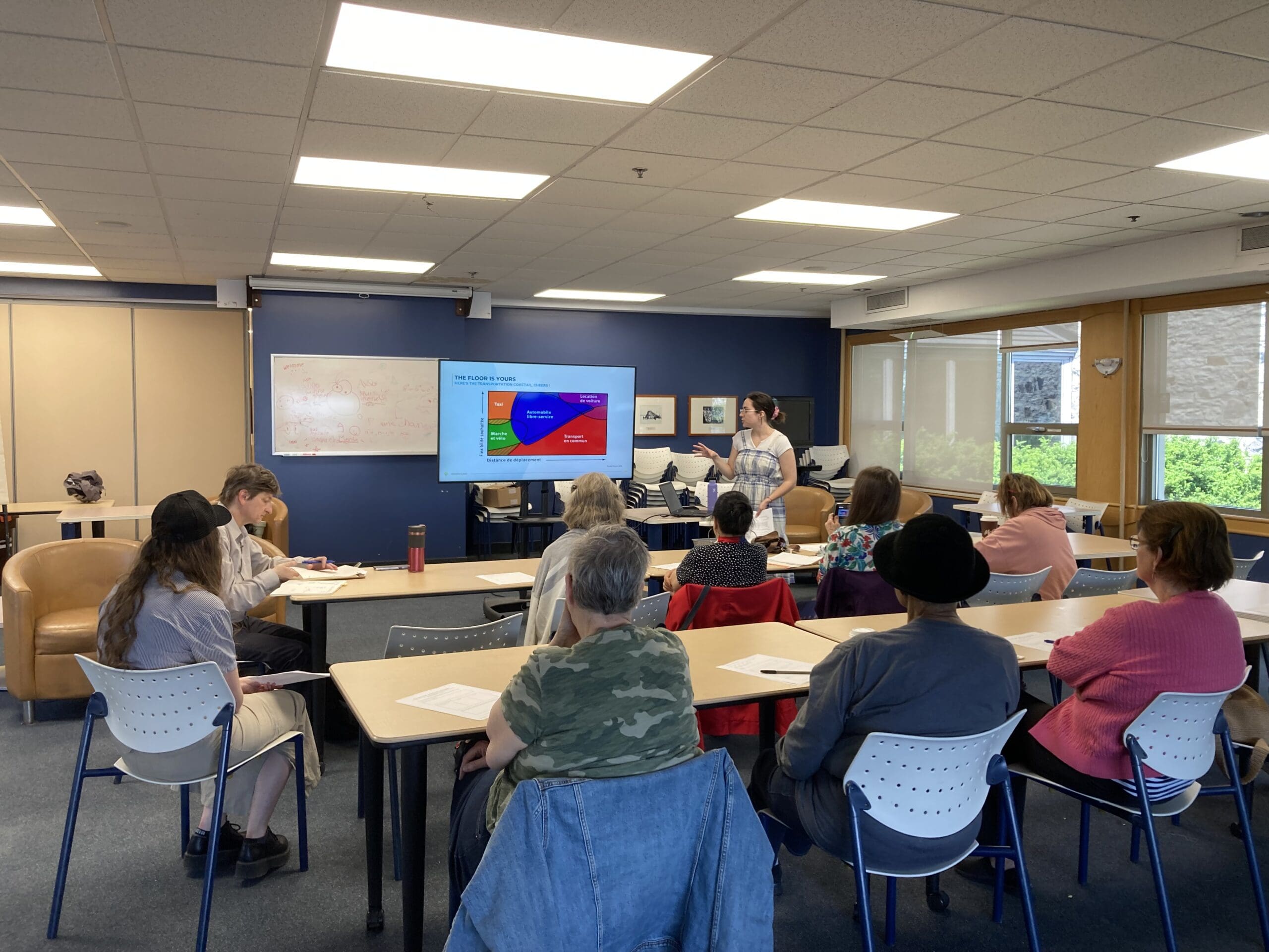 Un groupe de personnes est assis dans une salle de classe, écoutant attentivement un présentateur de MobilisAction Lachine. Le présentateur est debout près d'un écran affichant un diagramme circulaire coloré. Des tables et des chaises sont disposées en rangées, des notes sur certains bureaux, et de grandes fenêtres apportent de la lumière naturelle.