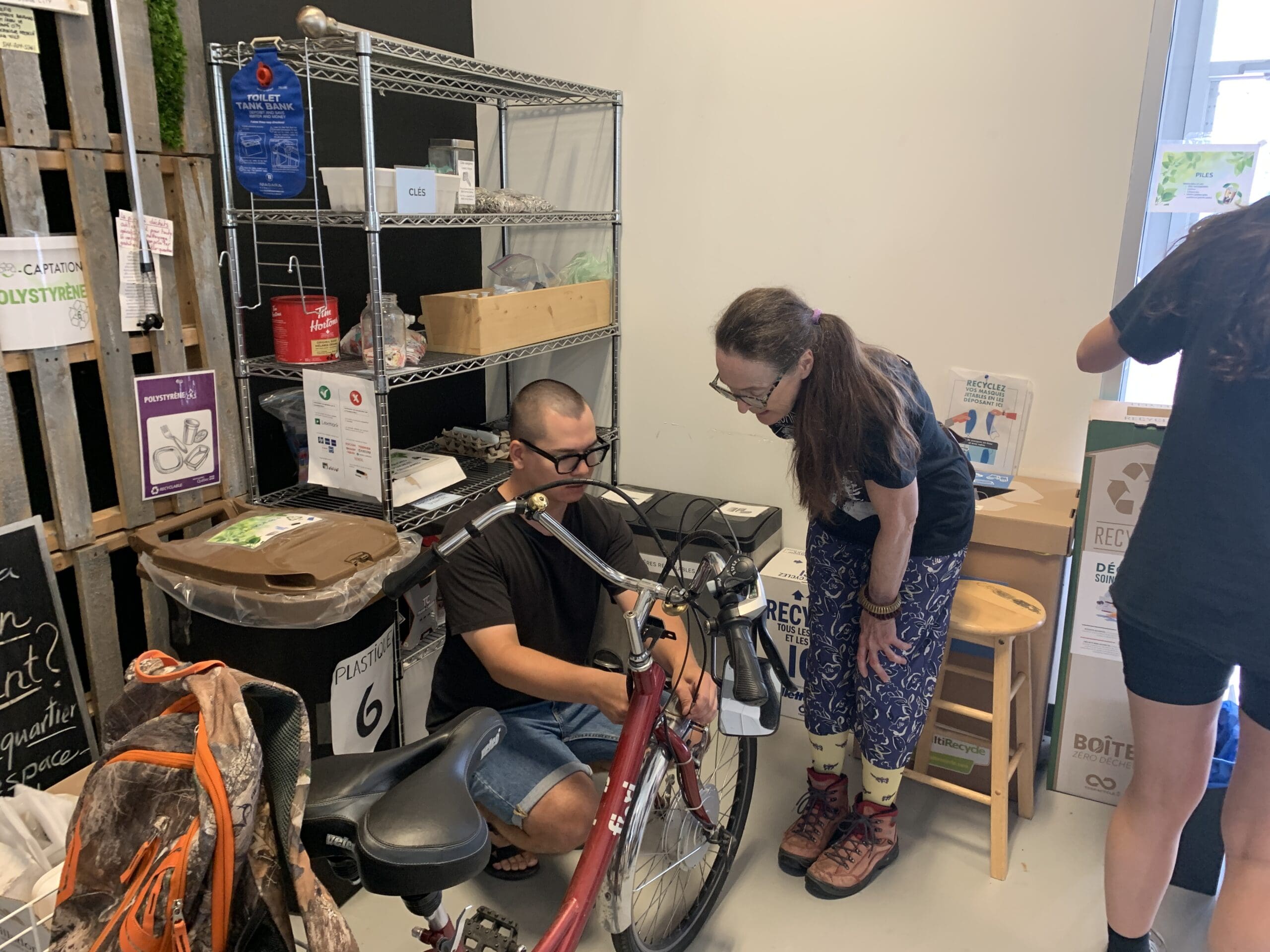 Dans une pièce encombrée d'étagères, de boîtes et de panneaux de recyclage, une personne portant des lunettes s'agenouille pour réparer un vélo rouge de MobilisAction Lachine, tandis qu'une autre se penche pour regarder. Pendant ce temps, une autre personne est partiellement visible sur la droite.