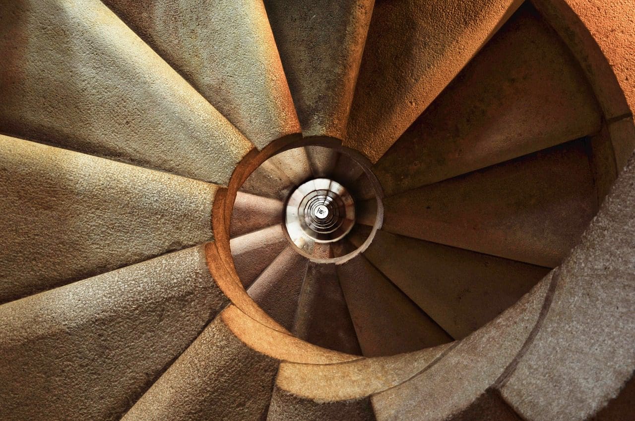 Un escalier de pierre en spirale vu du dessus, mettant en valeur son motif circulaire et ses tons bruns chauds. Les marches et l'axe central créent un dessin géométrique fascinant, la lumière et l'ombre accentuant la texture de la structure.