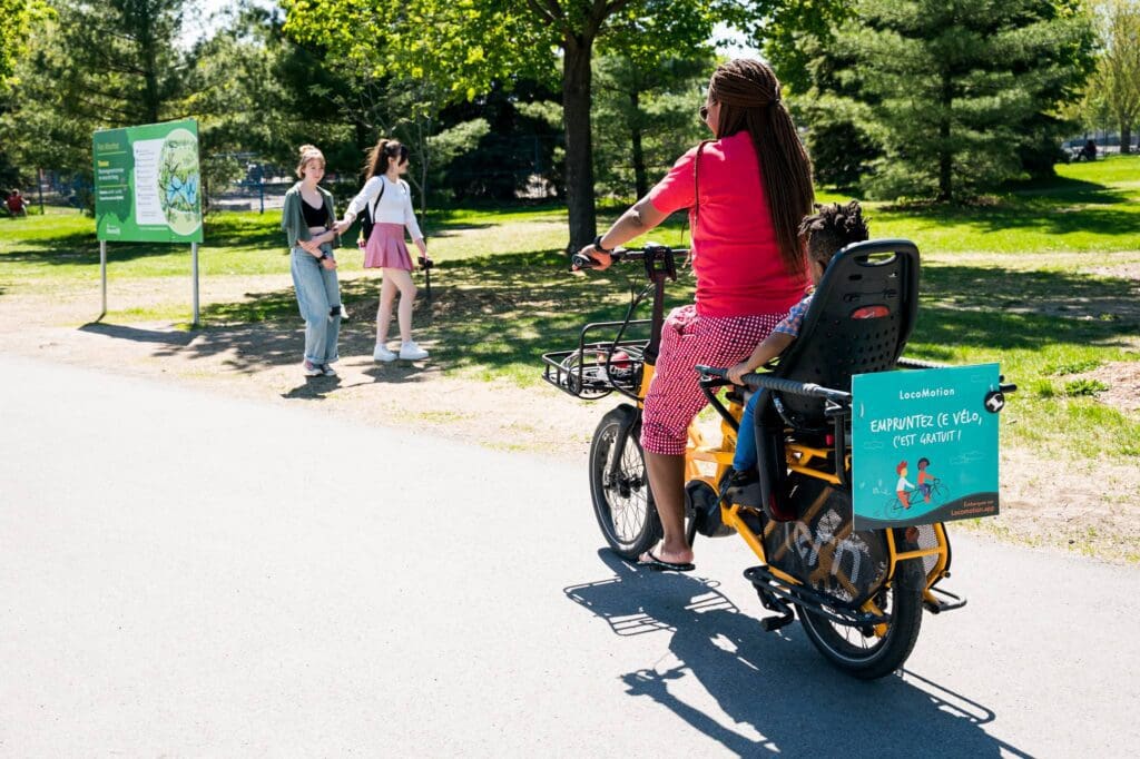 Une personne aux cheveux tressés roule à vélo sur un chemin avec un enfant dans un siège fixé à l'arrière. Deux personnes marchent à proximité. Le vélo porte un panneau de promotion du tourisme local. Journée ensoleillée avec des arbres et de l'herbe verte.