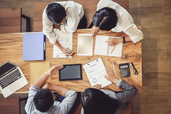 Quatre personnes sont assises à une table en bois et examinent des documents et des tablettes numériques. Un ordinateur portable, une calculatrice et des carnets de notes se trouvent également sur la table. Ils sont en train de discuter ou de planifier, des tableaux et des graphiques étant visibles parmi les documents.
