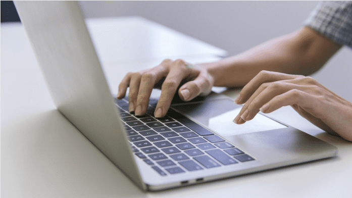 Une personne tape sur le clavier d'un ordinateur portable, les deux mains posées sur l'appareil. L'accent est mis sur les mains et le clavier, avec un bureau blanc en arrière-plan.