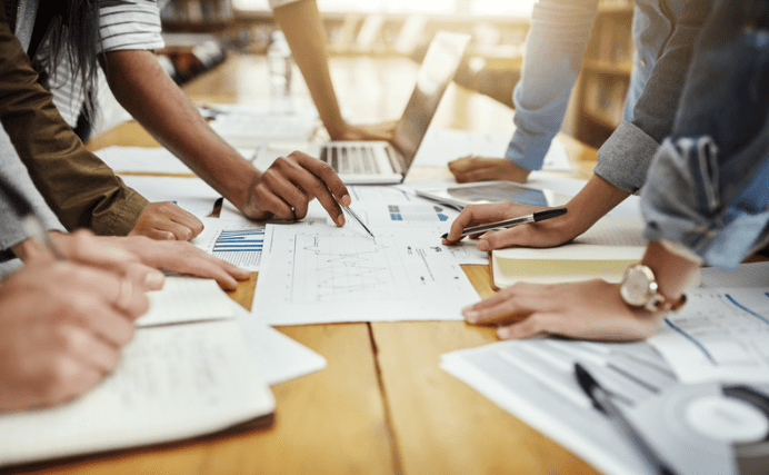 Un groupe de personnes autour d'une table, examinant et discutant de divers tableaux et graphiques imprimés sur papier. Une personne pointe un document à l'aide d'un stylo tandis que d'autres prennent des notes. Un ordinateur portable est également visible sur la table.