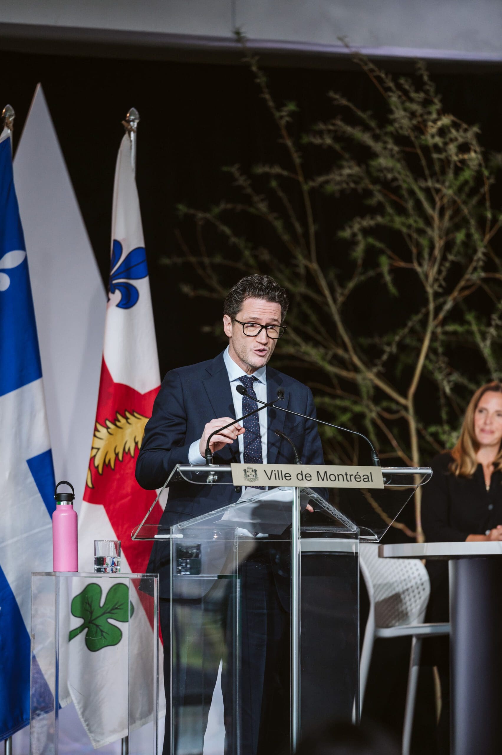 Benoit Charette, bien habillé, parle sur un podium étiqueté Ville de Montréal, encadré par des drapeaux et un arbre. Une bouteille d'eau rose est posée sur un support en verre à côté de lui, tandis qu'une femme est assise attentivement à la table derrière lui.