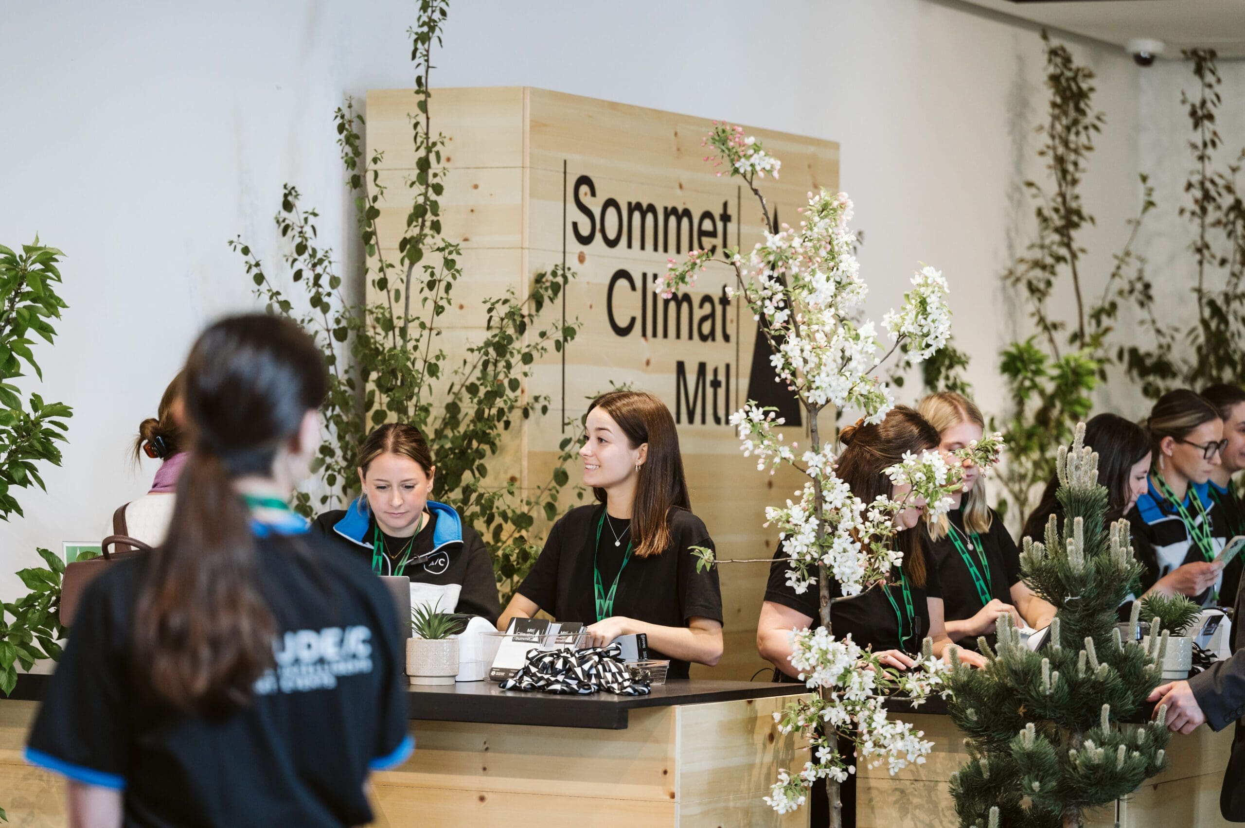 Entrée du Sommet Climat 2024. Des femmes distribuent les badges au participant, dans la bonne humeur. Derrière elles se trouve une installation en bois avec écrit "Sommet Climat Mtl". Il y a beaucoup de fleurs et de plantes autour.