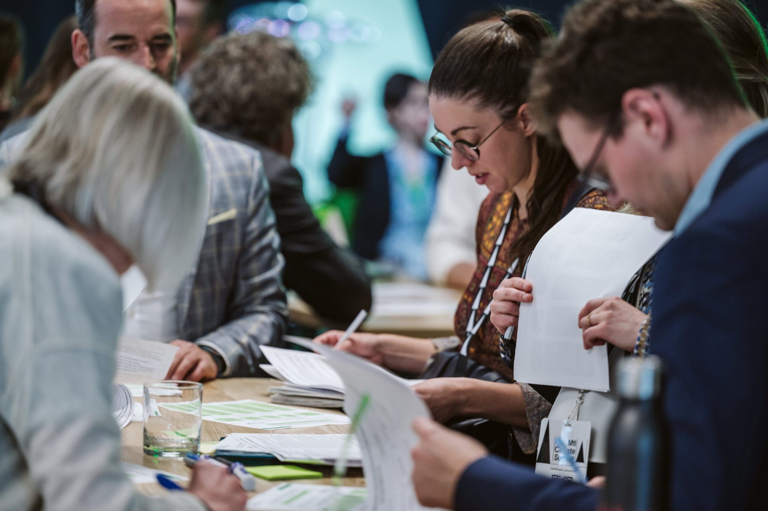Un groupe de personnes est assis à une table dans l'effervescence de la conférence Sommet Climat Montréal 2024, tenant des documents et prenant des notes. Les personnes semblent engagées et concentrées, l'arrière-plan flou suggérant une atmosphère animée.