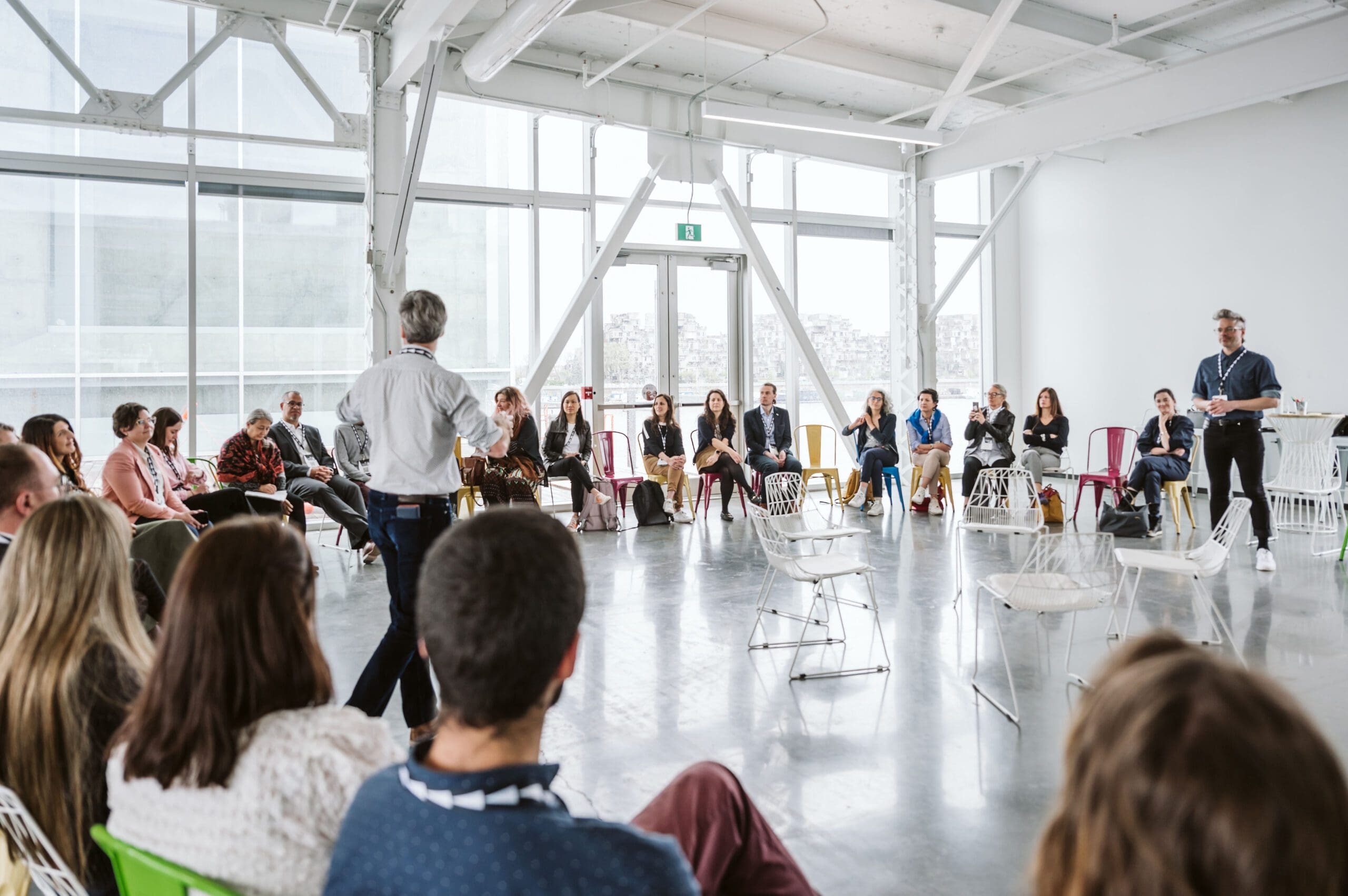 Dans une salle moderne et lumineuse dotée de grandes fenêtres, les participants au Sommet Climat Montréal 2024 sont assis en cercle. Alors qu'une personne s'adresse au groupe et qu'une autre se tient en face, leurs chaises - blanches et colorées - restent majoritairement occupées, reflétant l'engagement de chacun dans cette discussion dynamique.