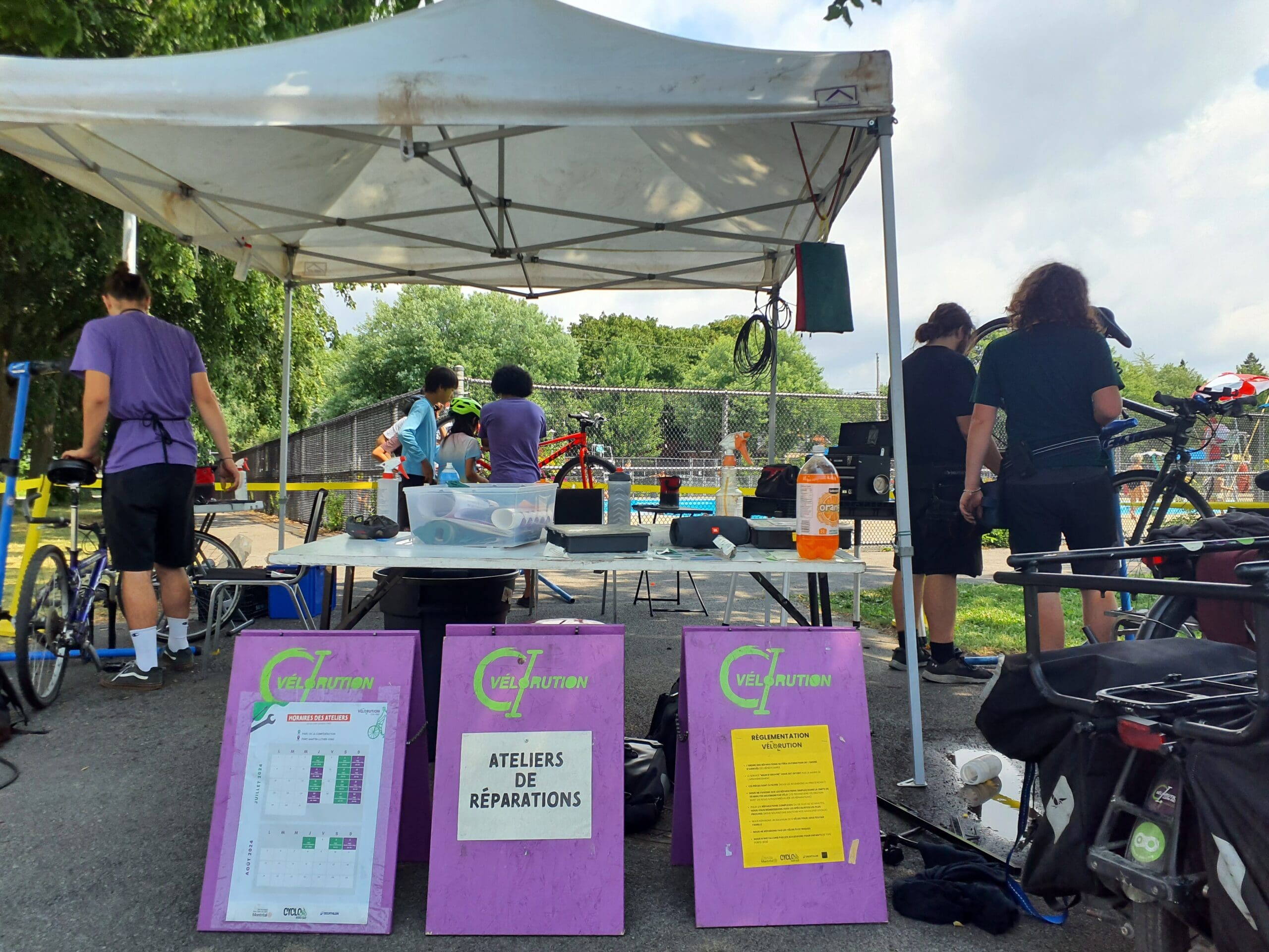 Des membres du projet Vélorution Couronne réparent des vélos sous une tente blanche lors d'un événement en plein air. Deux panneaux violets affichent en français des informations sur les ateliers de réparation. Des outils sont éparpillés sur la table, avec plusieurs vélos en arrière-plan.