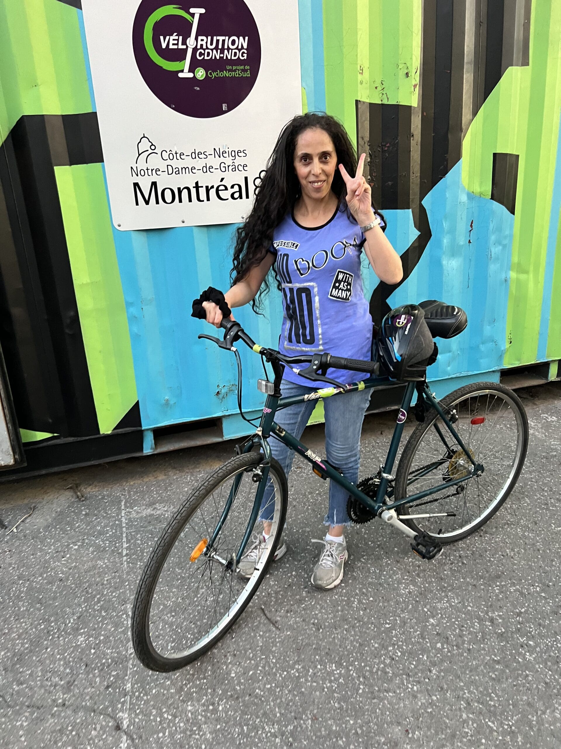 Une femme aux longs cheveux bouclés, vêtue d'une chemise mauve, se tient debout, tenant une bicyclette et affichant un signe de paix. Le mur coloré derrière elle affiche fièrement le projet Vélorution Couronne, Montréal, Côte-des-Neiges.