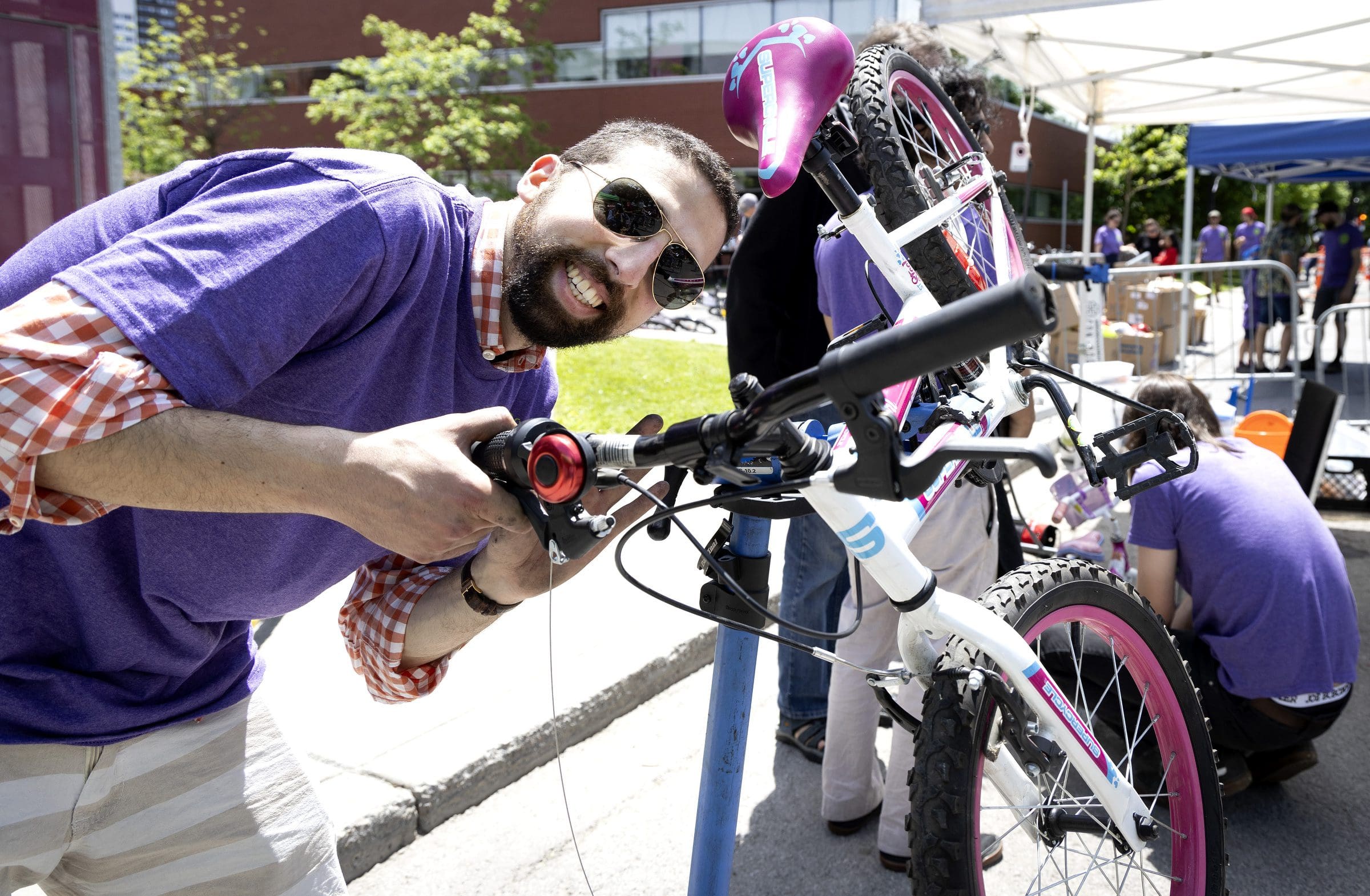 Un homme souriant portant des lunettes de soleil et une chemise violette répare un vélo rose et blanc dans le cadre du projet Vélorution Couronne. Une autre personne travaille sur un vélo à proximité, entourée d'arbres et d'un bâtiment en briques.