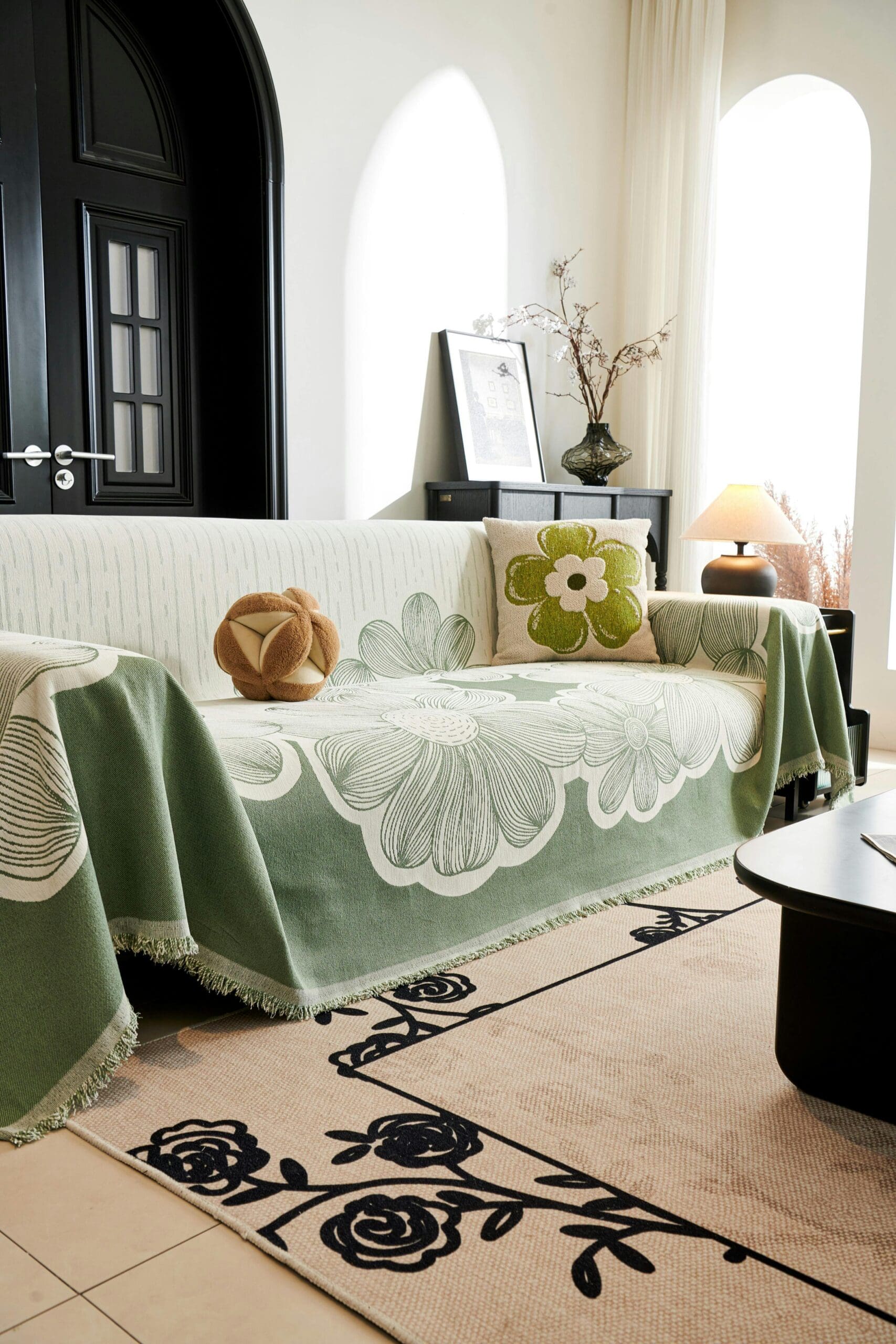 Un salon élégant présente un canapé drapé d'un jeté vert à motifs floraux. Un coussin orné d'une grande fleur est posé sur le canapé. Une table avec une lampe et des vases se trouve à l'arrière-plan. La pièce est dotée de fenêtres cintrées et d'un tapis à motifs floraux.