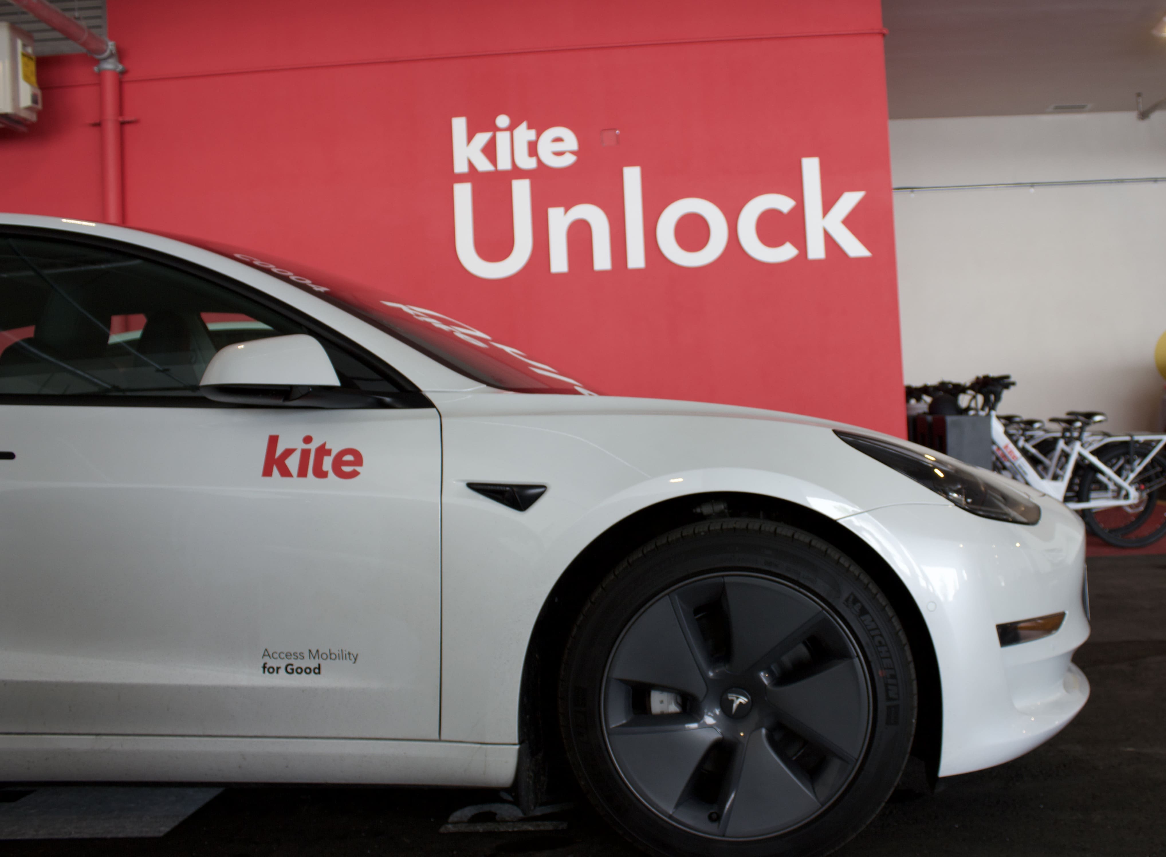 Un parking moderne avec un ensemble de voitures blanches. Les murs sont ornés de publicités colorées de Kite Mobility et du texte « Rideshare ». Des bornes de recharge pour véhicules électriques sont visibles, ce qui renforce l'ambiance futuriste et écologique.