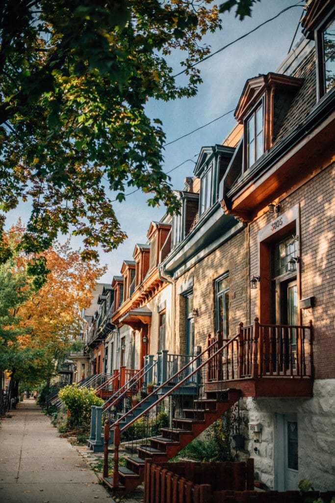 Une rangée de charmantes maisons de ville en brique de style ancien, aux toits pentus et aux lucarnes, borde une rue ombragée par des arbres. Les arbres arborent des couleurs d'automne et le ciel est clair, ajoutant une ambiance chaleureuse à la scène résidentielle.
